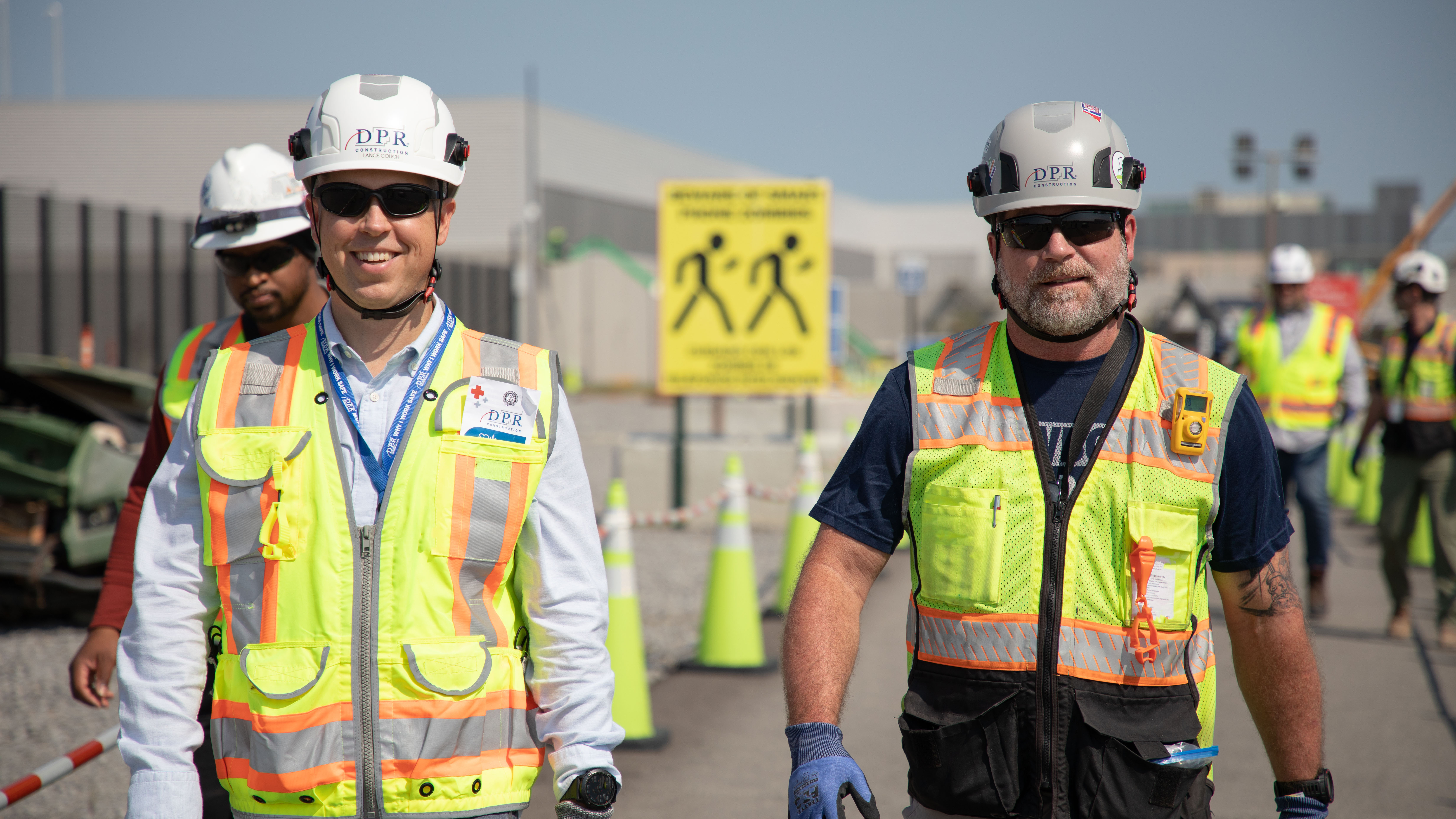 Two workers wearing PPE walk on a jobsite with other workers in the background.