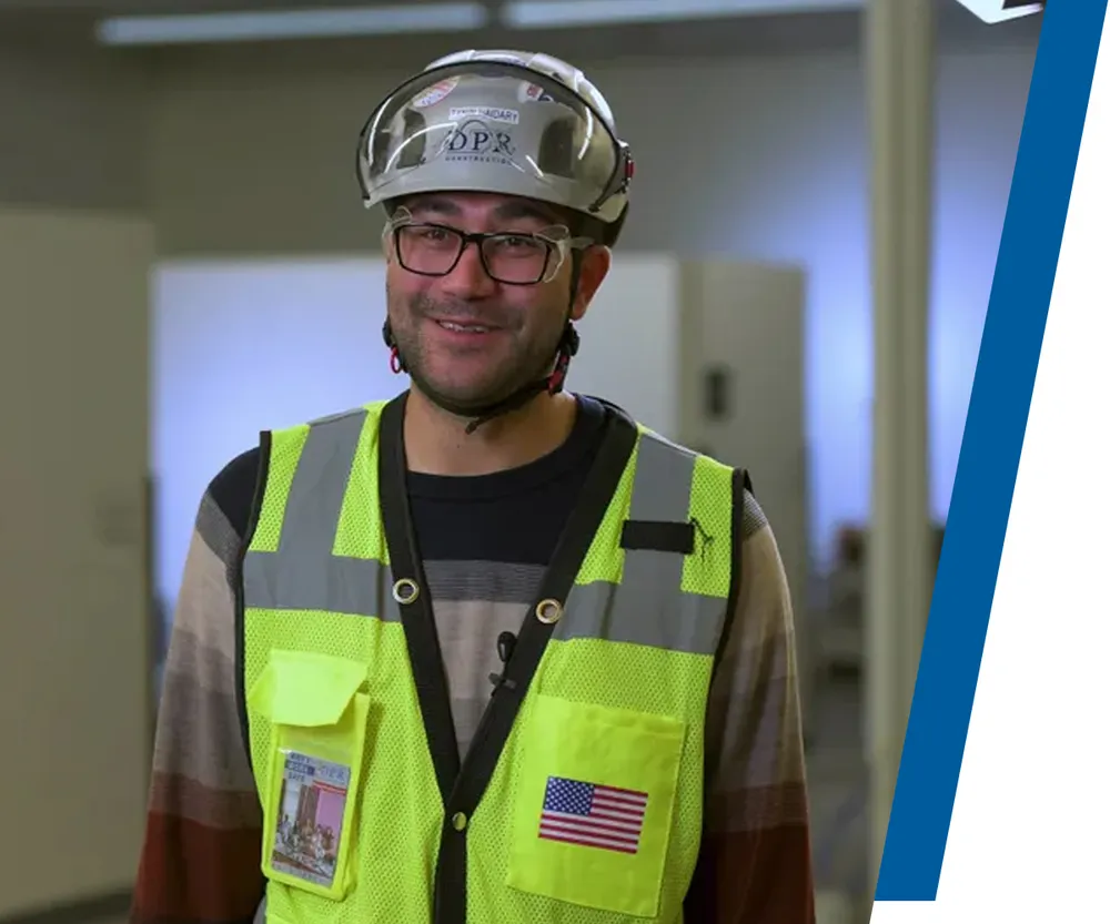 A builder wearing personal protective equipment smiles at the camera in an office trailer environment